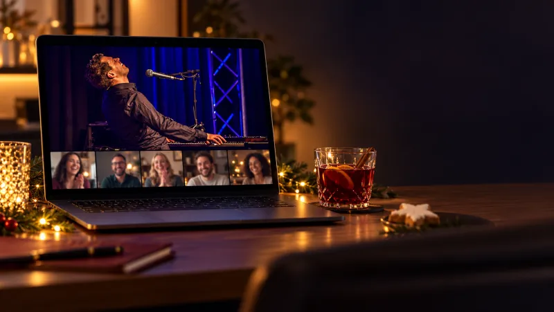 Laptop auf einem weihnachtlichen Schreibtisch, im Videocall spielt ein Musiker am Klavier, darunter lachende Kolleginnen und Kollegen